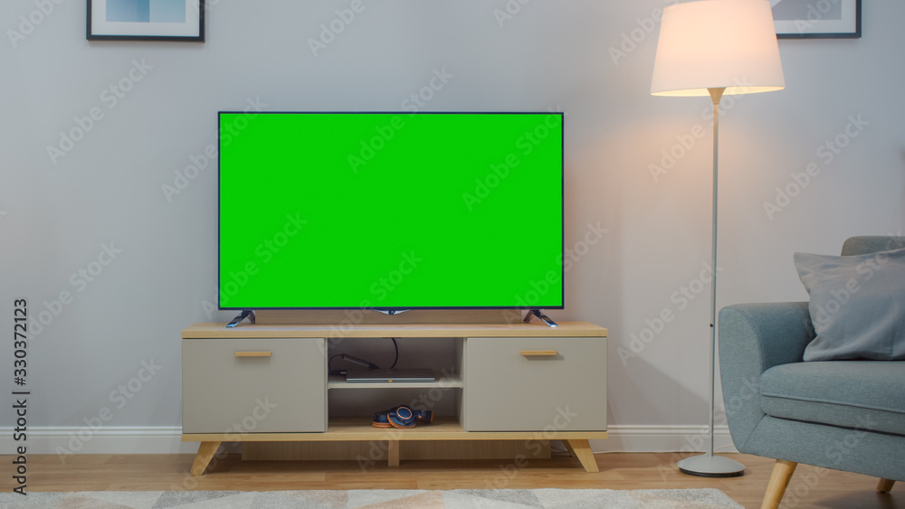 Shot of a TV with Horizontal Green Screen Mock Up. Cozy Living Room at Day Time with a Chair and Lamps Turned On at Home. Shot of a TV with Horizontal Green Screen Mock Up. Cozy Living Room at Day Time with a Chair and Lamps Turned On at Home.