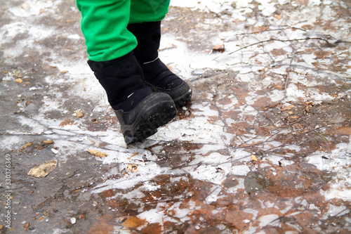 The boy breaks the thin ice in a puddle with his foot. The child knocks his boot on the white ice. Winter. December.