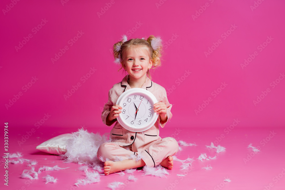 Studio shot of a contented, beautiful little girl posing in pink pajamas. Funny baby girl in pyjamas standing on a pink background with a pillow, feathers and a clock Studio shot of a contented, beautiful little girl posing in pink pajamas. Funny baby girl in pyjamas standing on a pink background with a pillow, feathers and a clock