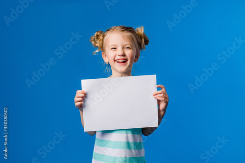 baby girl holding a white sheet.Cute little girl with a white sheet of paper.blue background.space for text.A little girl holds an empty piece of paper photo