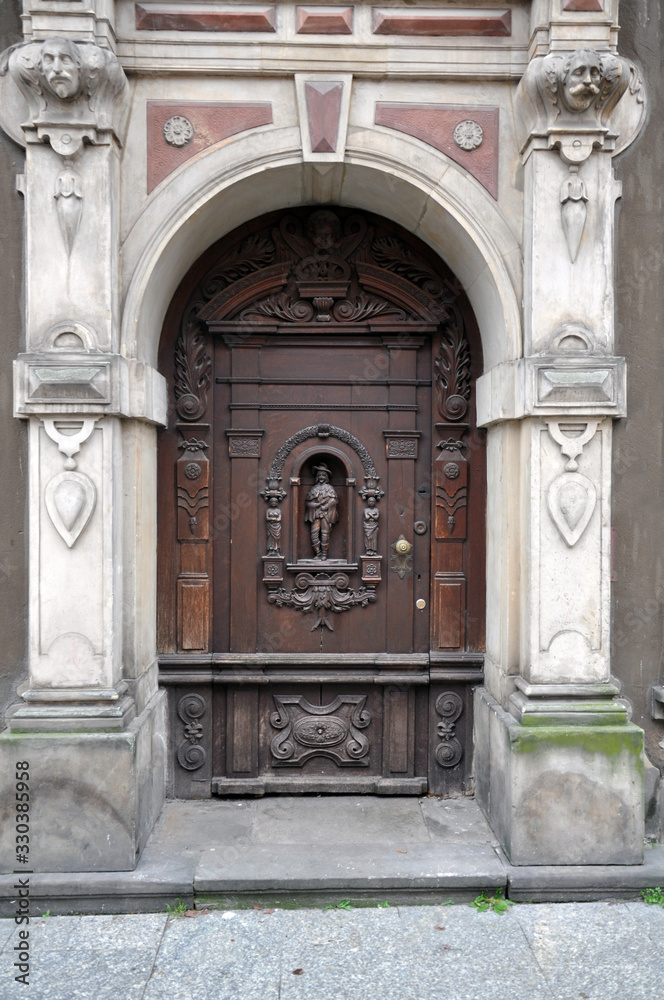 An old carved wooden door, with elements of forging, framed by sculptures. The facade is in the Baroque style. Poland, Gdansk 2019.