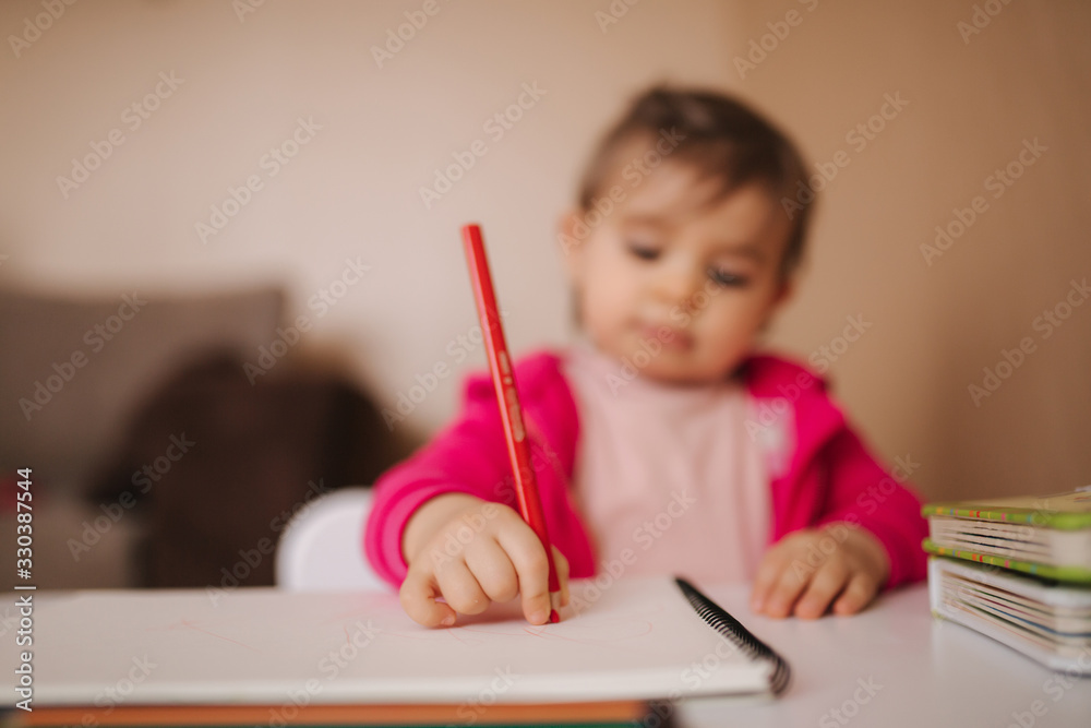 Happy baby girl sit at the table and wthire something. Little girl use pencil for drawing on white paper at home Happy baby girl sit at the table and wthire something. Little girl use pencil for drawing on white paper at home
