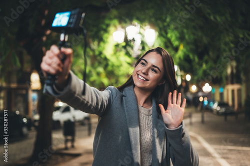 Female vlogger recording with digital camera showing hi. Smiling woman taking selfie video on light night city. Traveler making video for her blog. Vlogger uses photo camera for shoot social media photo