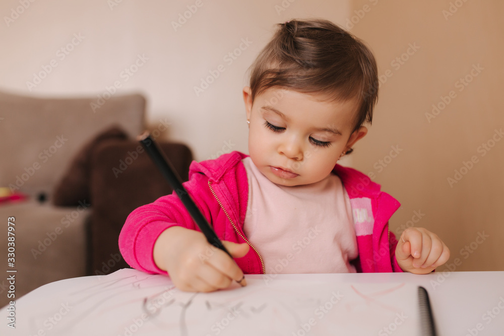 Happy baby girl sit at the table and wthire something. Little girl use pencil for drawing on white paper at home Happy baby girl sit at the table and wthire something. Little girl use pencil for drawing on white paper at home