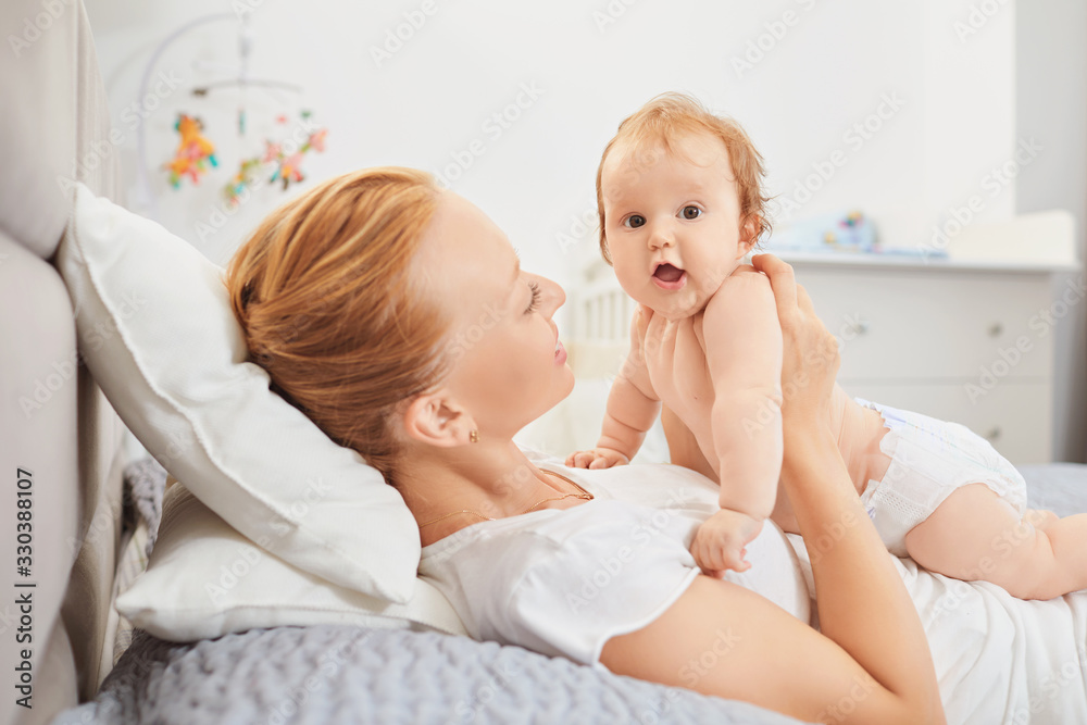 Happy mother smiles at baby lying on bed in room. Happy mother smiles at baby lying on bed in room.