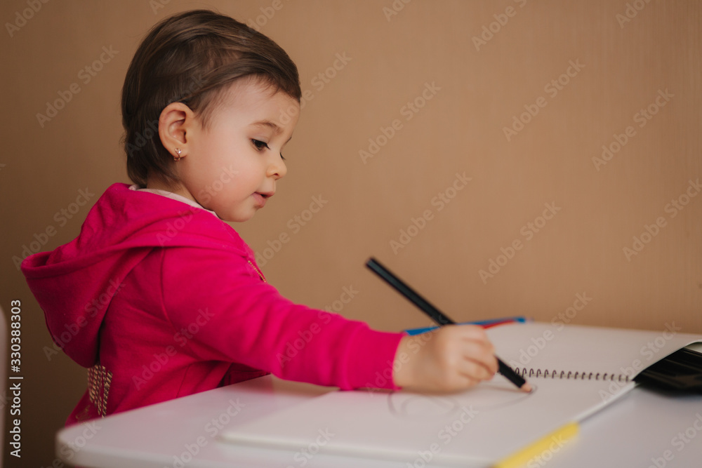 Happy baby girl sit at the table and wthire something. Little girl use pencil for drawing on white paper at home Happy baby girl sit at the table and wthire something. Little girl use pencil for drawing on white paper at home