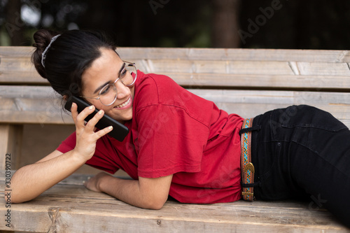 Young attractive woman talking through the phone and smiling in a park, sit down in a bench