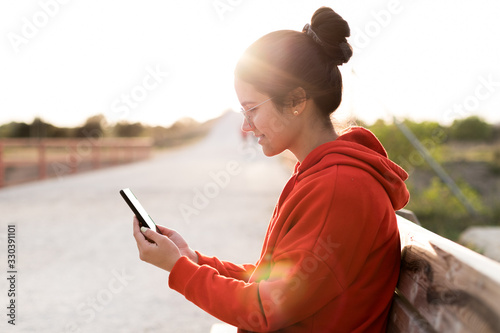 Young woman with glasses on her twenties chatting on her phone while sitting in a bench of a park. She is smiling and wearing a red sweater and a ponytail. She is backlit with daylight at sunset