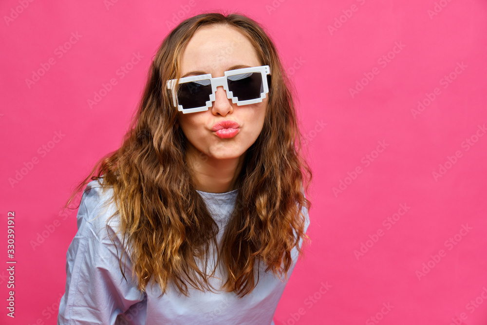 Stylish caucasian girl in a pale blue t-shirt and 8-bit glasses shows a kiss with lips on a pink background.