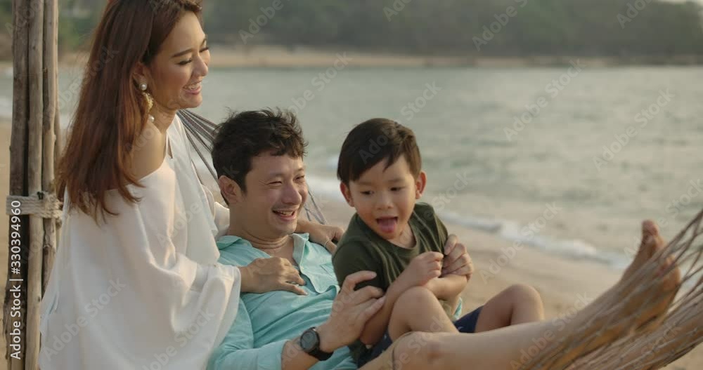 Lovely asian family enjoying in a beach hammock together. Slow motion shot of Young asian couple and little son lying in hammock together during summer holiday at the beach near the sea at sunset. Lovely asian family enjoying in a beach hammock together. Slow motion shot of Young asian couple and little son lying in hammock together during summer holiday at the beach near the sea at sunset.