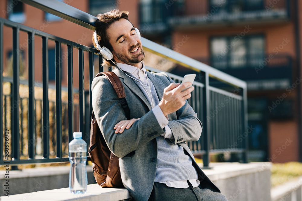 Naklejka premium Happy businessman with headphones using mobile phone outdoors.