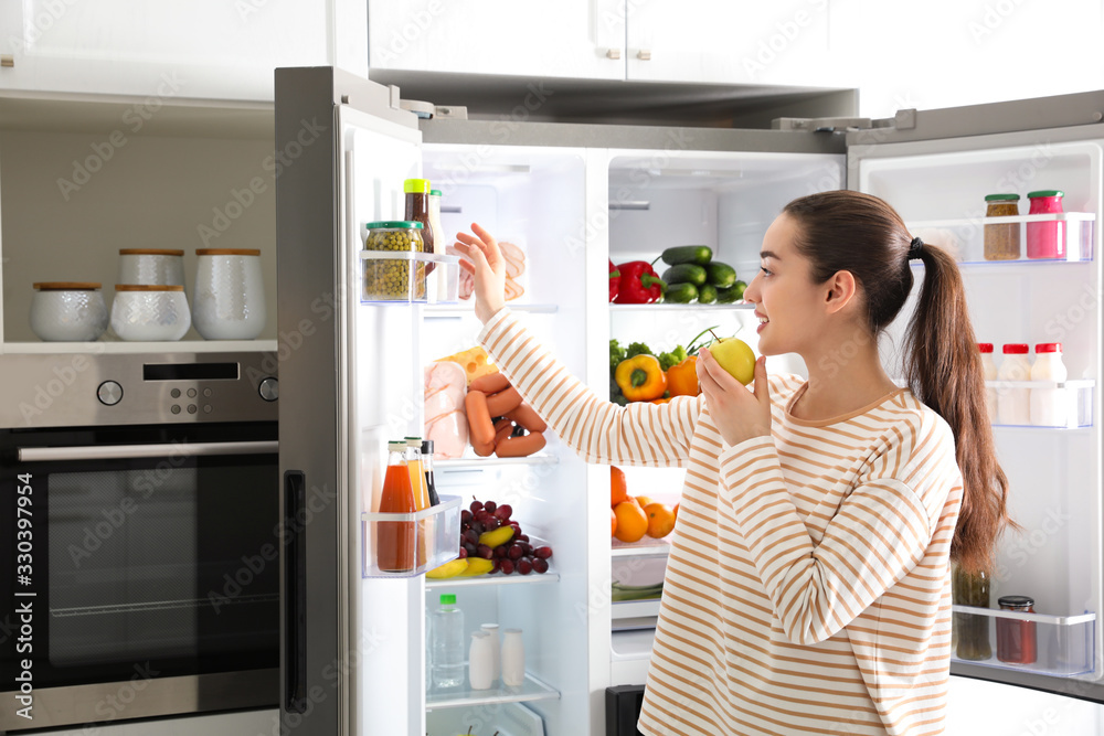 Young woman with apple near open refrigerator in kitchen Young woman with apple near open refrigerator in kitchen