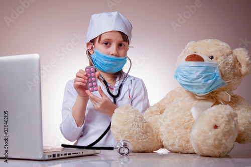 Little cute child girl doctor in protective face mask gives the tablets to patient Teddy bear. Virus, healthcare and medicine concept. Horizontal image.