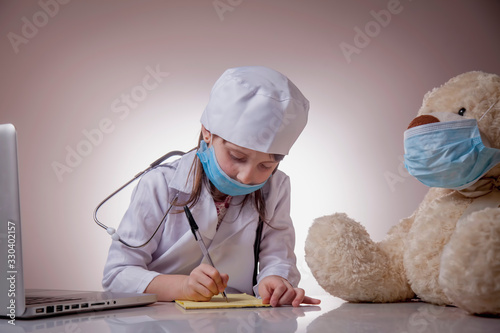 Little cute sad child girl doctor in protective face mask filling in paperwork of patient Teddy bear. Virus, healthcare and medicine concept.