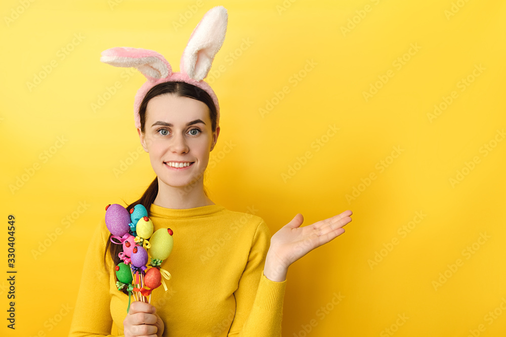 Studio shot of pleased woman wears bunny ears, raises palm as if showing something, gestures over blank space, holds colored eggs, dressed in sweater, models over yellow wall. Seasonal holiday concept