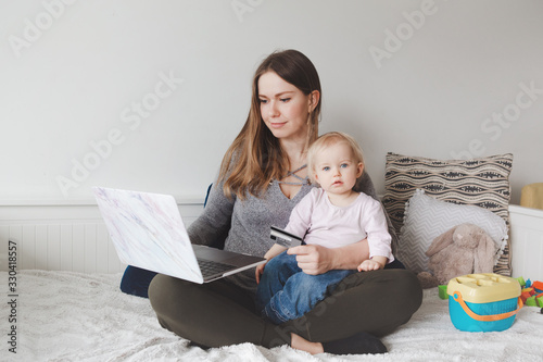 Young Caucasian mother with baby shopping online from home. Stay at home single mom with kid toddler daughter buying ordering products food on Internet and paying with a credit card. photo