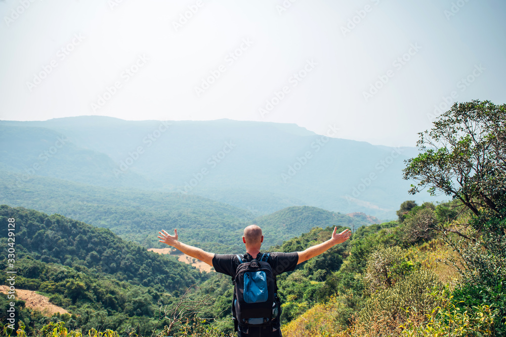 Man stands on top of hill and looks at the mountains covered with forest on sanny day. Brutal bald guy with a backpack spread his arms to the sides, view from the back. Concept of freedom and travel.
