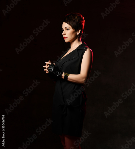 Sad woman in brutal black dress and leather gloves with cut fingers is standing sideways holding fingers interlocked