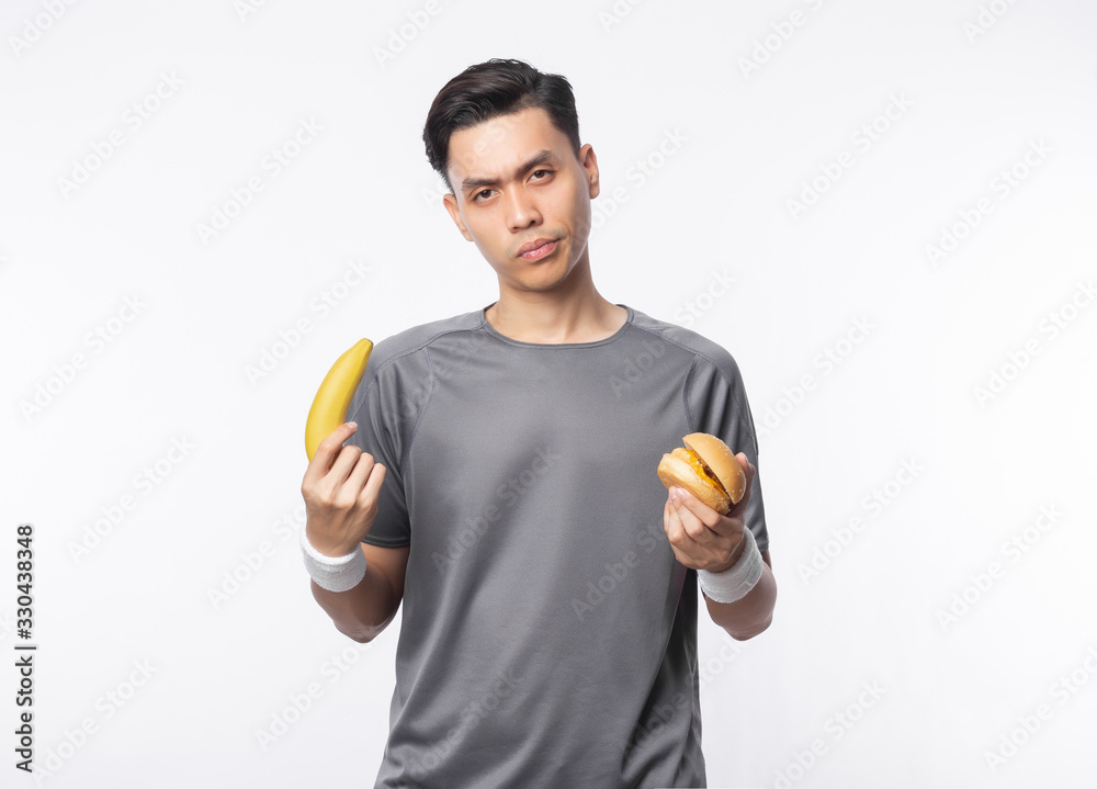 Young asian man in sport outfits holding banana and hamburger with doubts face isolated on white background. Young asian man in sport outfits holding banana and hamburger with doubts face isolated on white background.