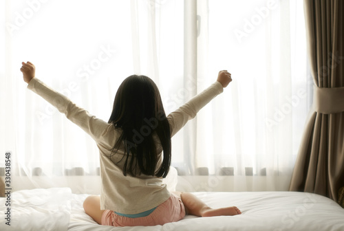 Asian child relax or kid girl wake up or woke up with stretch oneself after sleep for refreshing in morning on white bed and window curtain in bedroom at home or hotel on holiday and warm sunlight