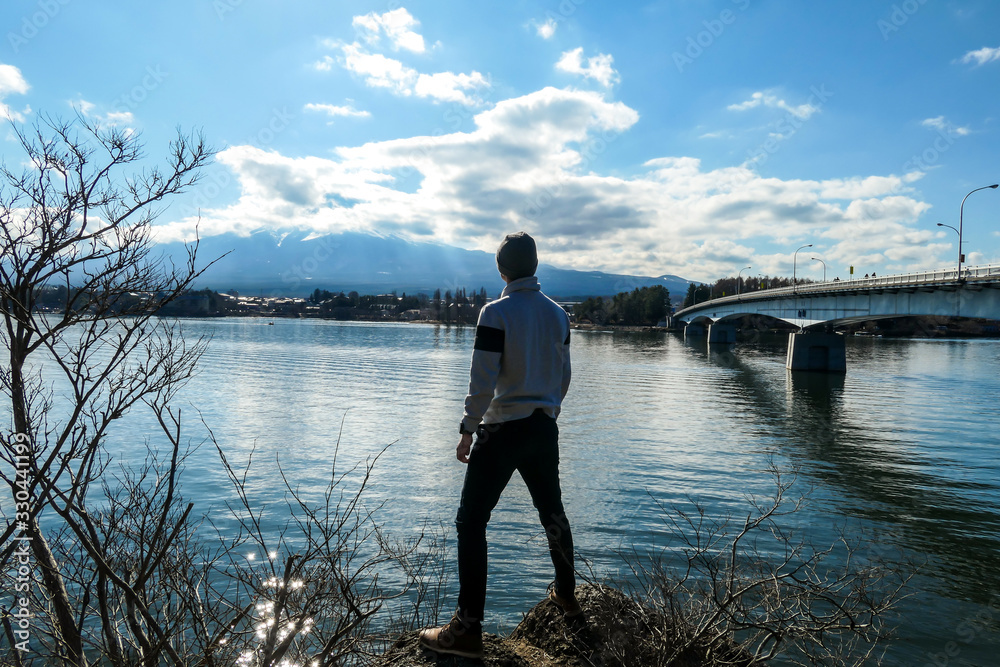 Man standing at the rocks, next to Kawaguchiko Lake, Japan with the view on Mt Fuji. The mountain surrounded by clouds. There is a bridge crossing the lake in the side. Serenity and calmness