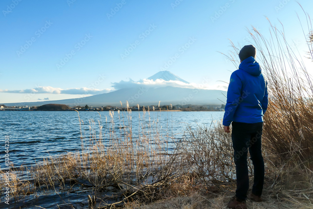 A man walking in between golden grass at the shore of Kawaguchiko Lake, Japan with the view on Mt Fuji. The man is enjoying the view on the volcano. The mountain surrounded by clouds. Serenity A man walking in between golden grass at the shore of Kawaguchiko Lake, Japan with the view on Mt Fuji. The man is enjoying the view on the volcano. The mountain surrounded by clouds. Serenity