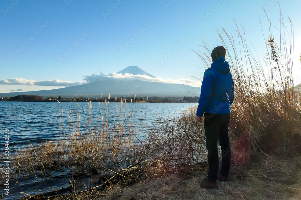 A man walking in between golden grass at the shore of Kawaguchiko Lake, Japan with the view on Mt Fuji. The man is enjoying the view on the volcano. The mountain surrounded by clouds. Serenity