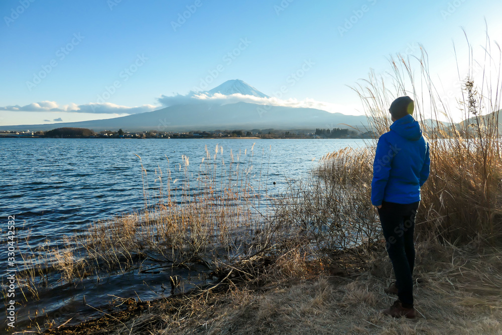 A man walking in between golden grass at the shore of Kawaguchiko Lake, Japan with the view on Mt Fuji. The man is enjoying the view on the volcano. The mountain surrounded by clouds. Serenity