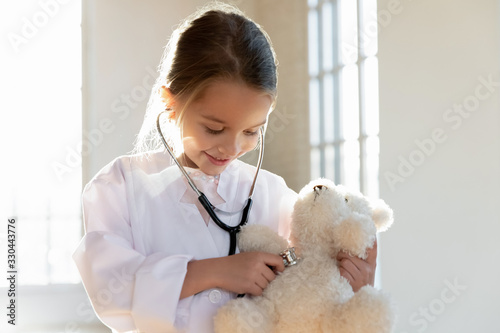 Fototapeta Naklejka Na Ścianę i Meble -  Head shot smiling adorable small girl wearing white coat, using stethoscope on teddy bear. Attractive little preschooler in uniform playing doctor patient game in clinic, children healthcare concept.