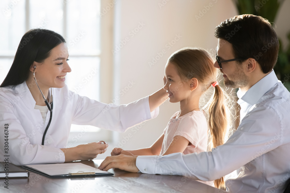 Smiling pleasant female pediatrician stroking head of preschool girl sitting on father s lap. Smiling doctor getting acquainted with small patient before starting checkup listening heartbeat lungs. Smiling pleasant female pediatrician stroking head of preschool girl sitting on father s lap. Smiling doctor getting acquainted with small patient before starting checkup listening heartbeat lungs.