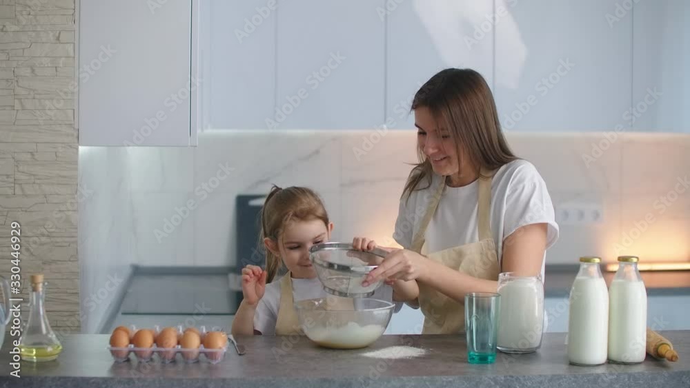 Mother and daughter sowing flour through the sieve into a bowl. Mother teaches daughter to knead a dough. Focus on the daughter. Mother and daughter sowing flour through the sieve into a bowl. Mother teaches daughter to knead a dough. Focus on the daughter.