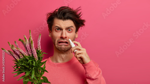 Indoor shot of frustrated man has watery red eyes, runny stuffy nose, uses drops, suffers from hay fever, has allergy on blooming plant, poses on pink background. Spring rhinitis, reaction on trigger photo