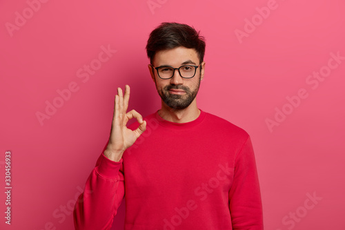 No problem concept. Bearded man makes okay gesture, has everything under control, all fine gesture, wears spectacles and jumper, poses against pink background, says I got this, guarantees something photo