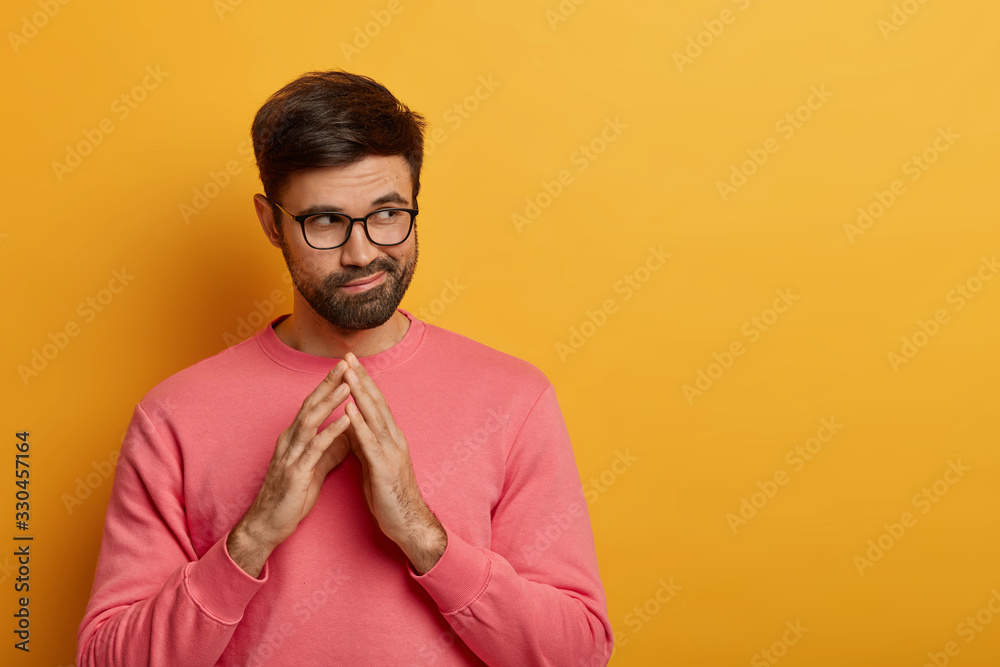 Tricky thoughtful bearded man steepls fingers, has some intention in mind, considers plans for future, looks aside mysteriously, has scheme, wears rosy sweater, isolated over yellow background
