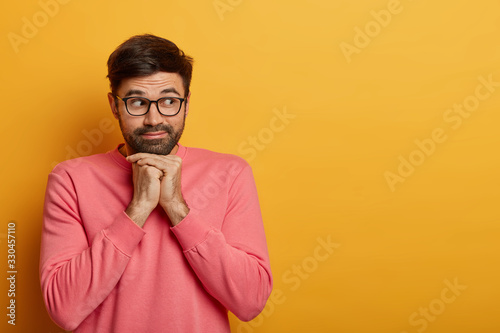 Handsome curious man looks right on empty yellow space, keeps hands under chin, considers buying something, has interest, wears spectacles and rosy jumper, poses indoor, anticipates event happen