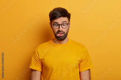 Portrait of bearded guy shows grimace, crosses eyes and sticks out tongue, plays around, going crazy, wears spectacles, everyday t shirt, poses against yellow background. Human face expressions © wayhome.studio 