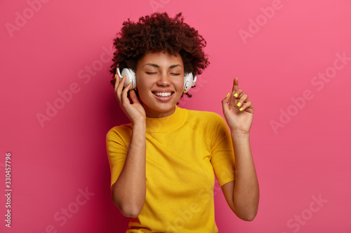Optimistic dark skinned woman closes eyes and smiles ecstatic, enjoys good sound quality, wears headphones for listening music, dances carefree, dressed in yellow clothes, isolated on pink wall