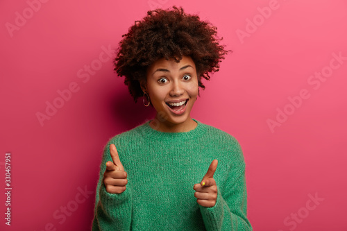 Pleased dark skinned female makes finger gun pistol, indicates at you, expresses choice, smiles pleasantly, wears green jumper, isolated on pink background, smiles upbeat, congratulates colleague