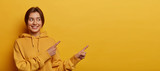 Smiling dark haired woman gives advice and points right, promots product, wears casual sweatshirt, poses against yellow background, indicates at advertisement, introduces promo with pleased expression