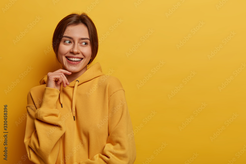 Pleasant looking Caucasian woman keeps hand on face, looks away with satisfaction, notices something pleasant, wears casual hoodie, isolated on yellow background, blank space aside for your promo Pleasant looking Caucasian woman keeps hand on face, looks away with satisfaction, notices something pleasant, wears casual hoodie, isolated on yellow background, blank space aside for your promo