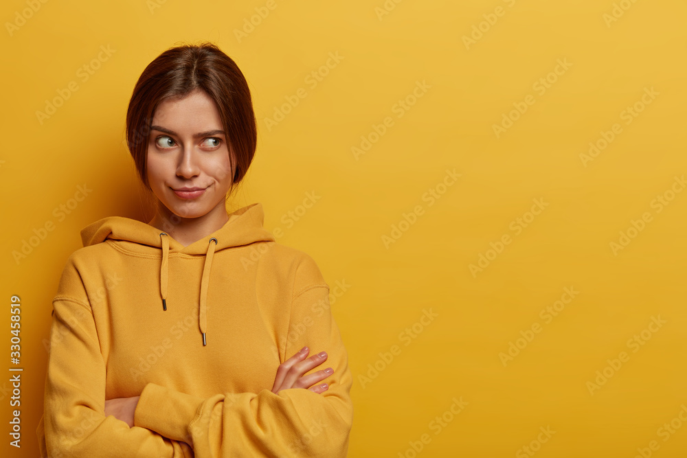 Fototapeta premium Horizontal shot of serious looking European woman crosses hands over chest, thinks about something with doubt, wears casual hoodie, poses against yellow studio wall, doesnt want to talk to you