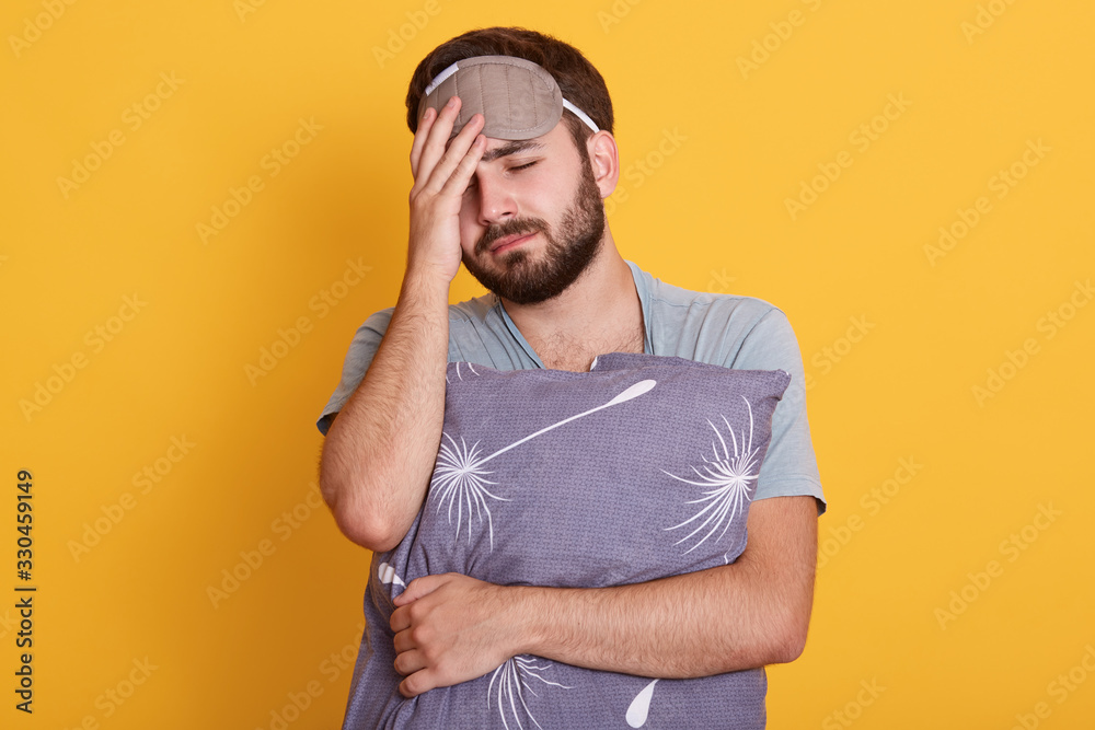 Studio shot of young unshaven male embracing gary pillow and holding hand on his forehead having terrible headache, suffering from pain, having sleepy tired facial expression,keeps eyes closed. Studio shot of young unshaven male embracing gary pillow and holding hand on his forehead having terrible headache, suffering from pain, having sleepy tired facial expression,keeps eyes closed.