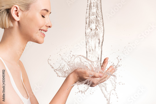 Photo of  young woman with clean skin and splash of water. Smiling woman with drops of water near her face. Spa treatment. Girl washing hands with water. Water and body. Water falling on human hands