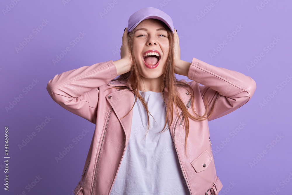 Attarctive woman in leather pink jacket covering ears and feeling irritation from loud repairing works, expressing dislike, looking directly at camera, screaming something. People emotions concept. Attarctive woman in leather pink jacket covering ears and feeling irritation from loud repairing works, expressing dislike, looking directly at camera, screaming something. People emotions concept.