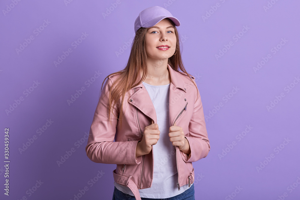 Studio portrait of young beautiful fashionable model wearing pink leather jacket, white shirt and cap. Women fashion and lifestyle concept. Confident female poses isolated over lilac studio background Studio portrait of young beautiful fashionable model wearing pink leather jacket, white shirt and cap. Women fashion and lifestyle concept. Confident female poses isolated over lilac studio background