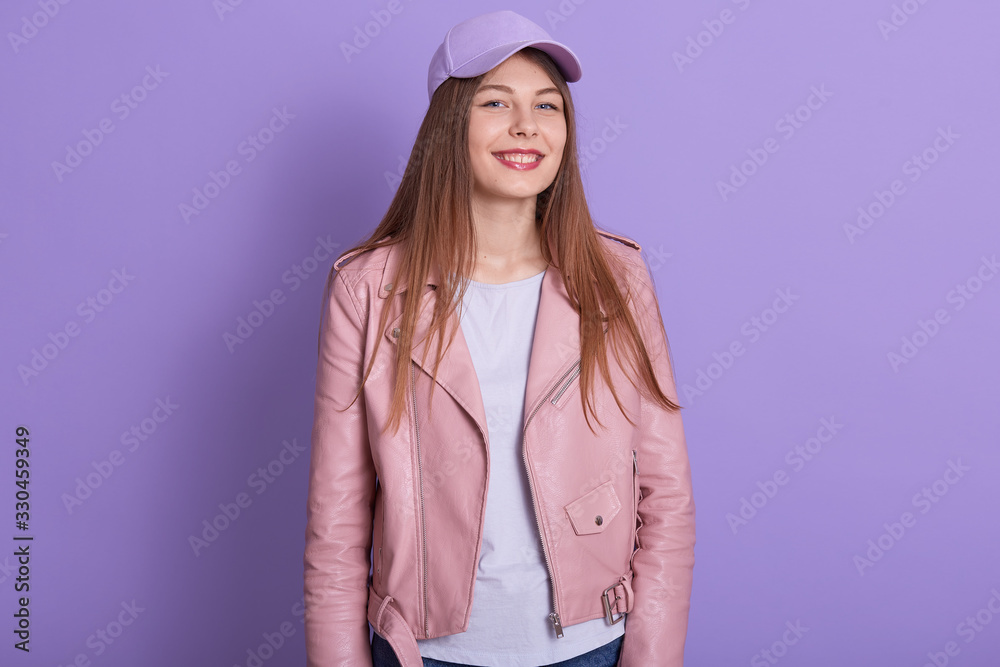 Closeup portrait of woman looking playful at camera, beautiful girl wearing white shirt, pink jacket and cap. Lady standing isolated over lilac studio background. Girl with pleasant facial expression. Closeup portrait of woman looking playful at camera, beautiful girl wearing white shirt, pink jacket and cap. Lady standing isolated over lilac studio background. Girl with pleasant facial expression.