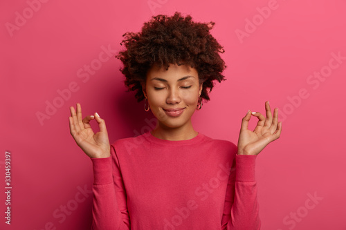 Relaxed satisfied patient curly woman makes yoga gesture, stands in lotus pose, keeps eyes closed, tries to forget about all problems, breathes deeply, wears pink casual jumper, models indoor