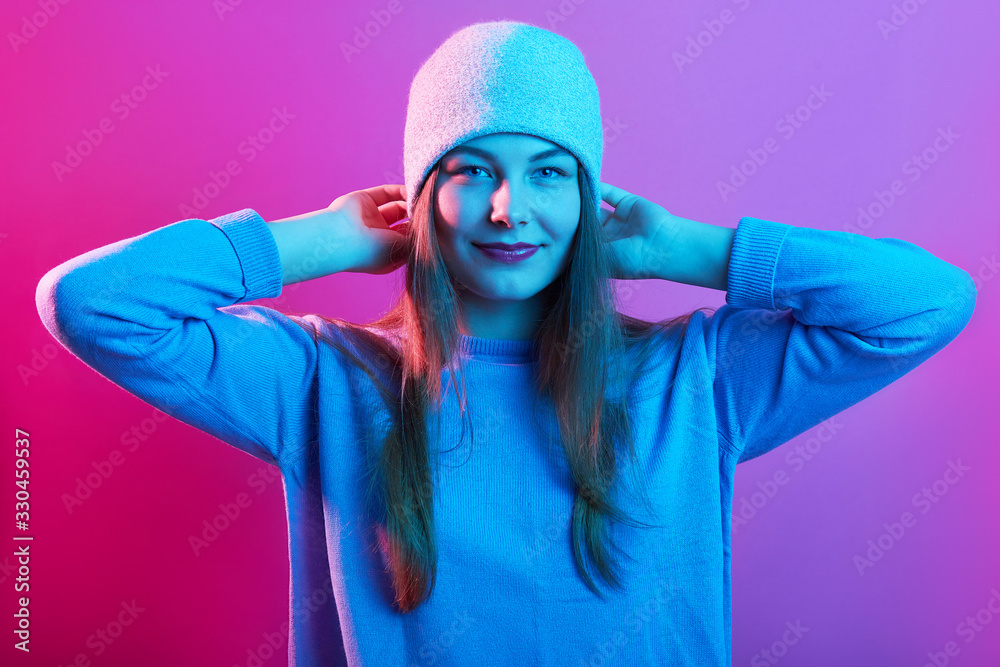 Picture of young adorable girl wearing sweater and cap, standing isolated over pink neon background, looking at camera with charming happy smile, keeping her hands behind his head, expresses happyness Picture of young adorable girl wearing sweater and cap, standing isolated over pink neon background, looking at camera with charming happy smile, keeping her hands behind his head, expresses happyness