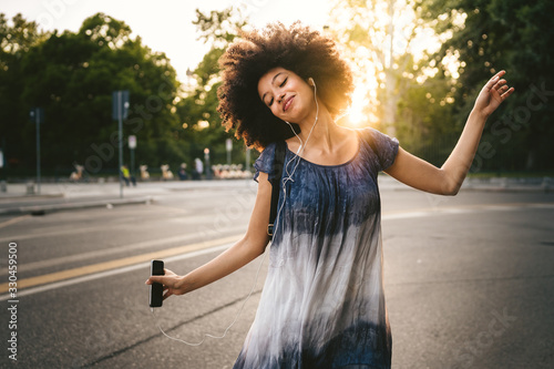Young woman dancing listening to the music with earphones from the smart phone outside a park at sunset on a summer day
