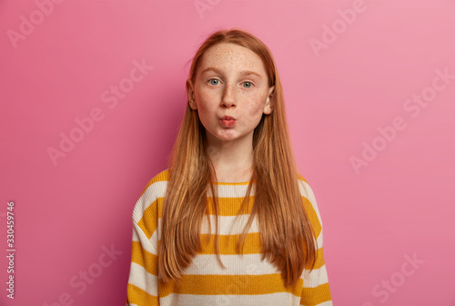 Slika na platnu Lovely red head teenage girl keeps lips folded, sends mwah, wants to kiss her mommy, wears striped casual sweater, looks with eyes full of love, isolated on pink backgroound